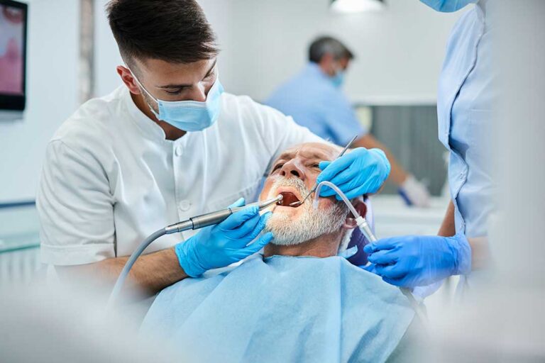 Dental hygienist cleaning a patient's teeth with instruments while the patient reclines in a dental chair.